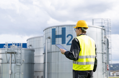 Engineer in a yellow hard hat and reflective vest holding a tablet in front of a hydrogen storage tank