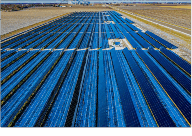Aerial view of a large solar farm with rows of photovoltaic panels stretching across open land near a rural highway.