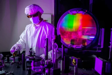 A scientist in protective gear works with high-powered laser equipment in a laboratory, surrounded by precision instruments and a large, colorful optical lens used for inertial fusion energy research.
