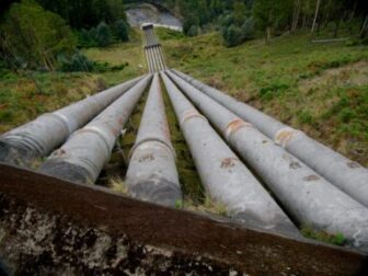Large industrial pipelines running down a hillside toward a river in a forested valley.
