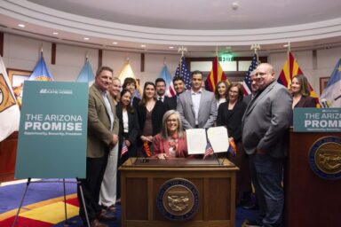Governor Katie Hobbs sits at her desk surrounded by officials and supporters as she signs Executive Order 2025-13, part of “The Arizona Promise” initiative focused on energy, opportunity, and economic growth.