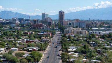 Aerial view of downtown Tucson, Arizona, with city buildings, green neighborhoods, and distant mountain ranges under a clear blue sky.