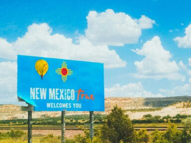 Welcome sign reading “New Mexico True Welcomes You” with desert landscape and blue sky in the background.